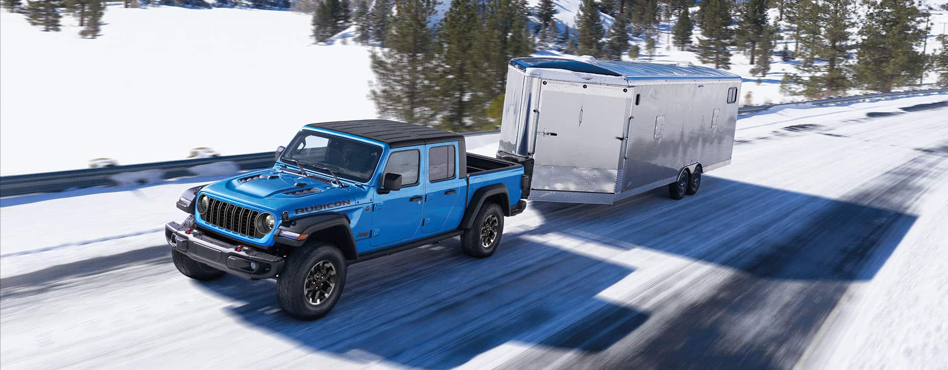 A blue 2024 Jeep Gladiator Rubicon being driven down a snow-covered highway as it tows an enclosed trailer.