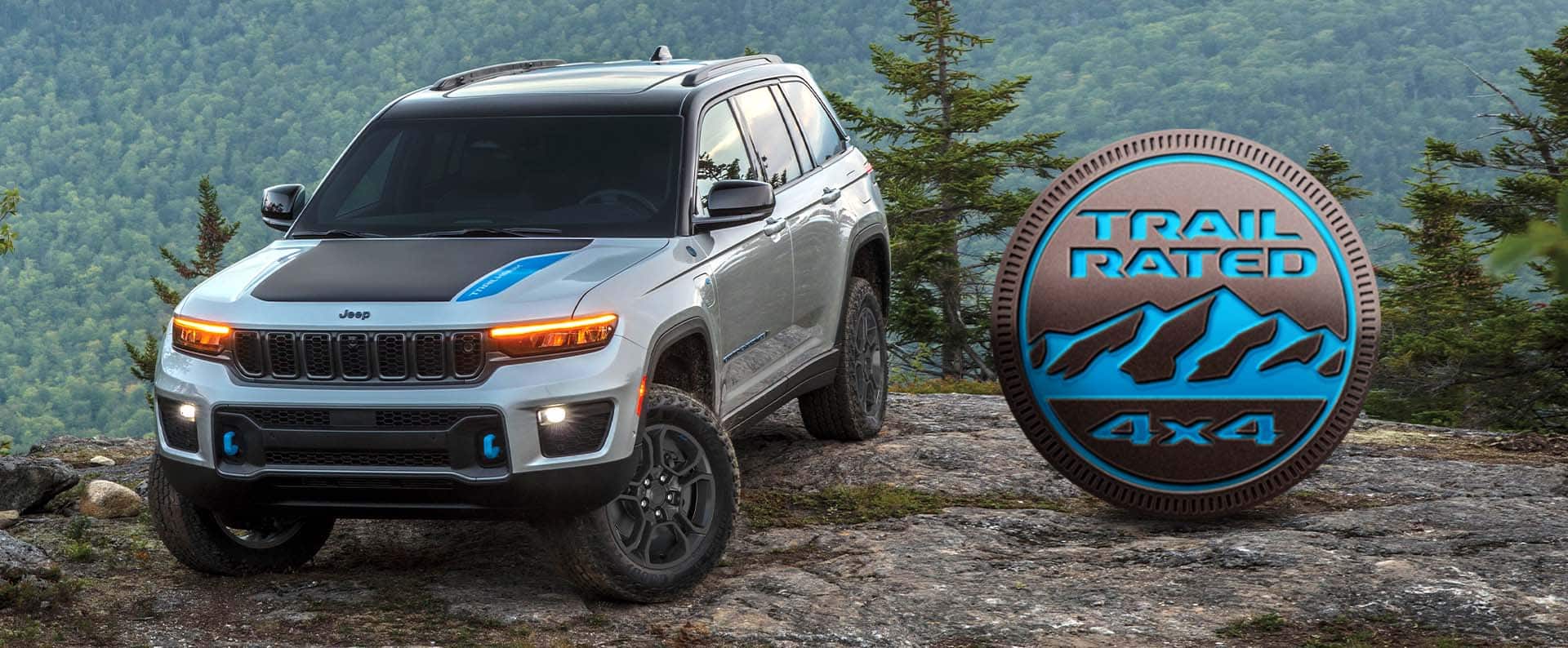 Un Jeep Grand Cherokee Trailhawk 4xe 2024 plateado estacionado en un claro fuera de la carretera, con un bosque de montaña de fondo. El emblema Trail Rated 4x4.
