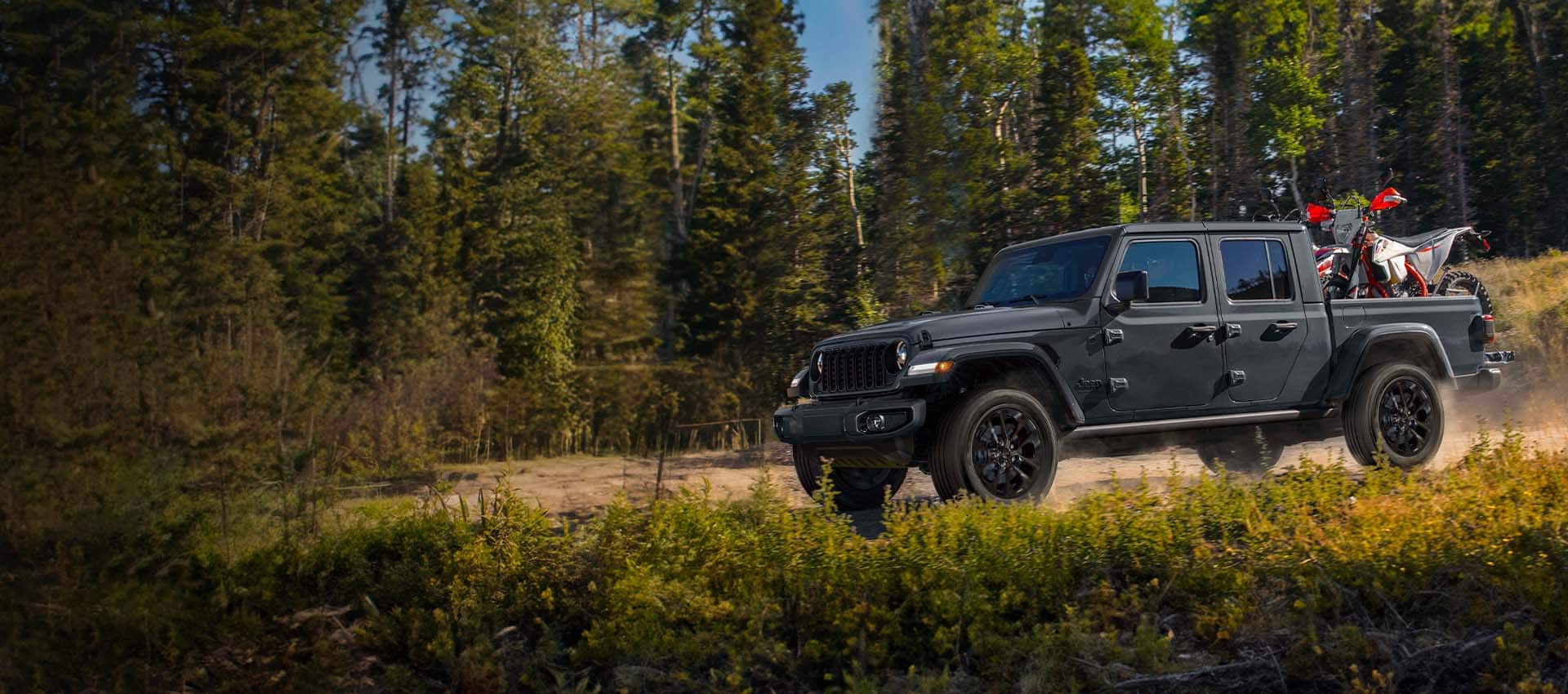 A black 2024 Jeep Gladiator Nighthawk being driven on a trail off-road in the woods with an ATV in its pickup bed.