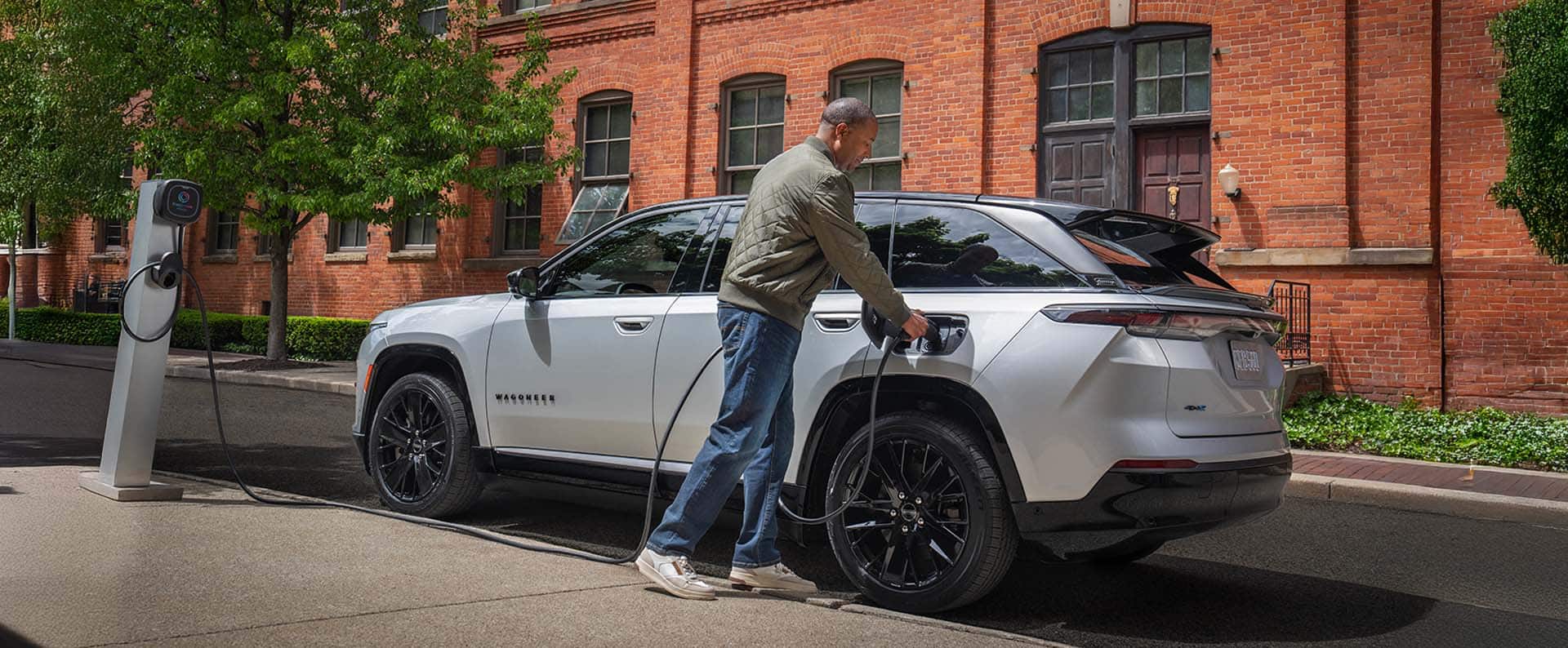 Un Jeep Wagoneer S Launch Edition 2024 plateado estacionado al lado de un edificio de ladrillo, con un hombre enchufando el cable de carga de una estación de carga pública en el puerto de carga del vehículo.